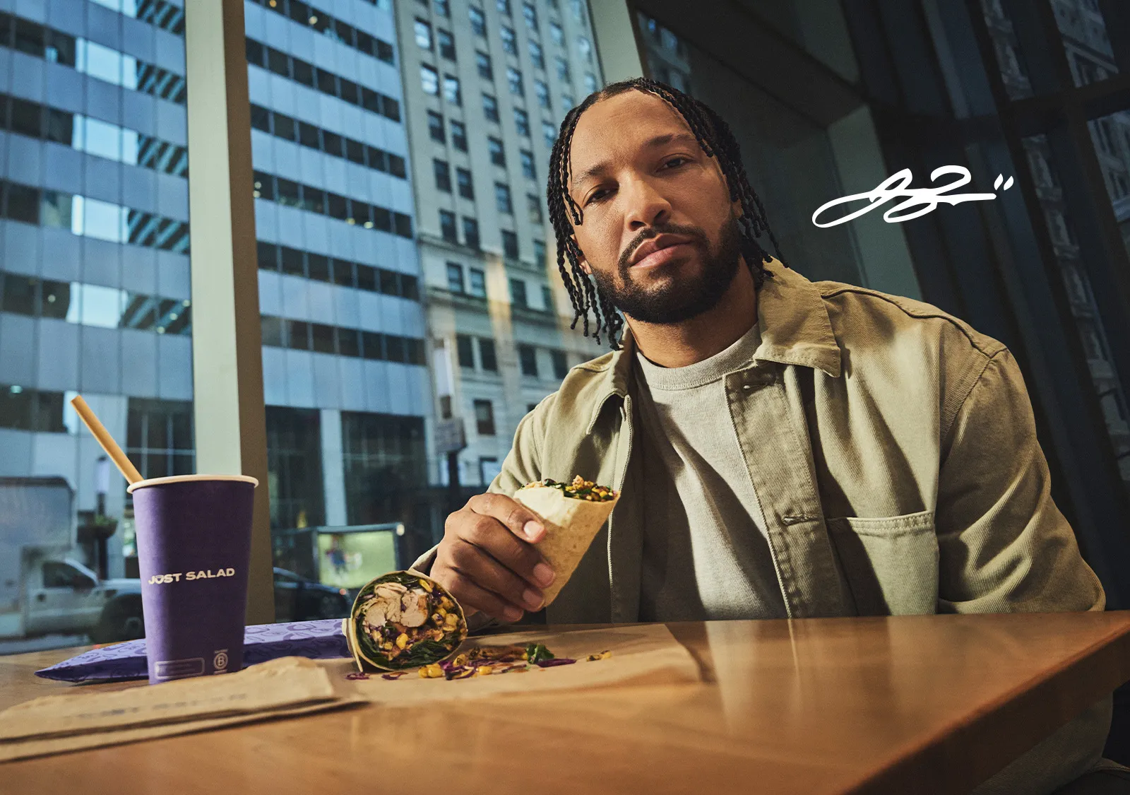 Jalen Brunson at a table by a window holding the Clutch Wrap, with a Just Salad cup beside him.